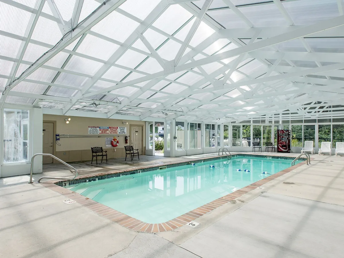 This image shows a bright, inviting indoor swimming pool with a transparent roof allowing natural light to flood the space. Lounge chairs line the poolside, and safety equipment is visible, emphasizing comfort and family-friendly amenities.