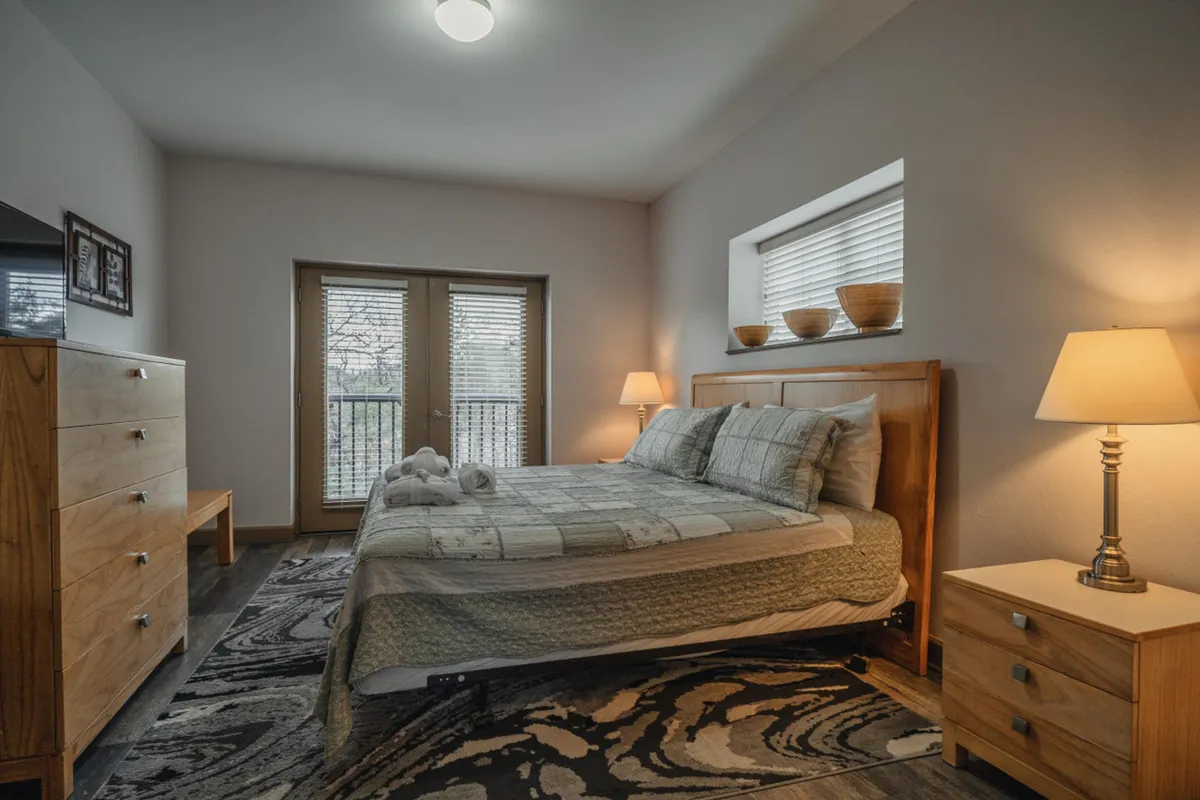 A cozy bedroom with a bed, dresser, sliding doors, lamp, and a soft rug. Natural sunlight filters into the room, creating a warm and inviting atmosphere perfect for relaxation and restful sleep.
