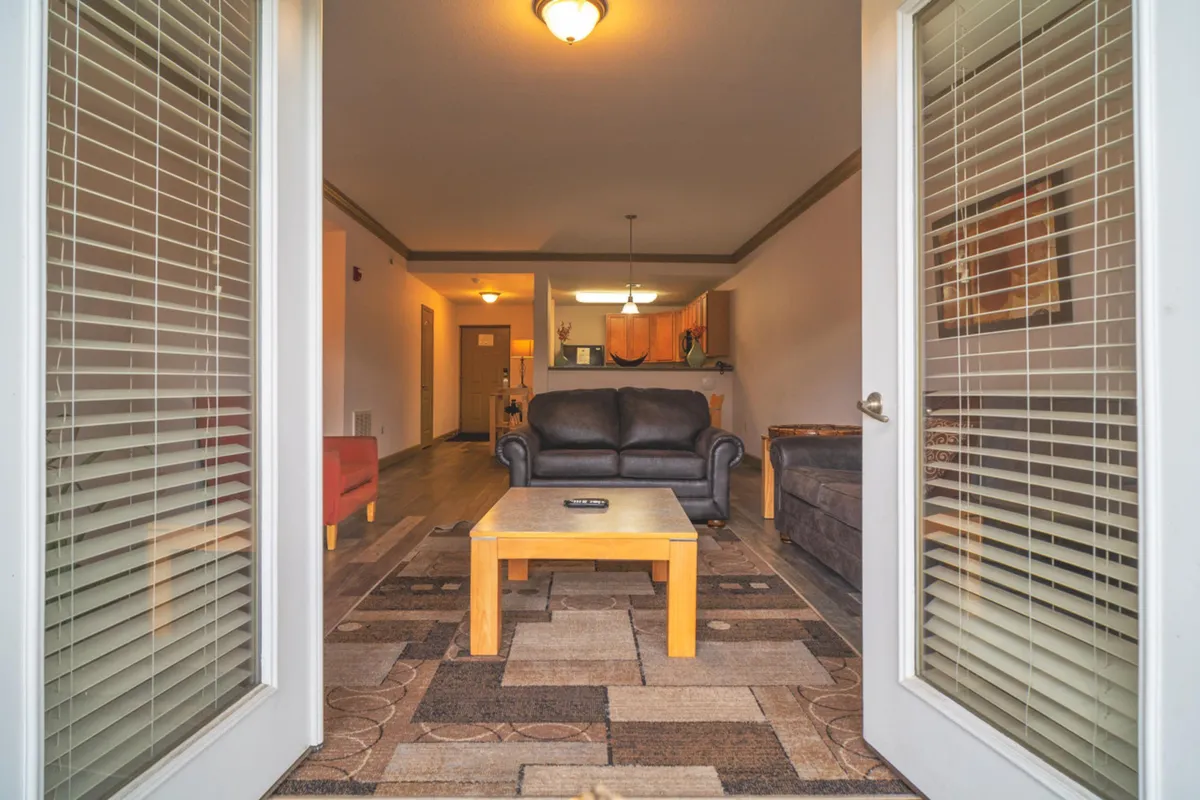 A bright, comfortable living area featuring a black leather sofa, a wooden coffee table, decorative rug, and a view into the kitchen area, suggesting a sociable and relaxed environment for spending time indoors.