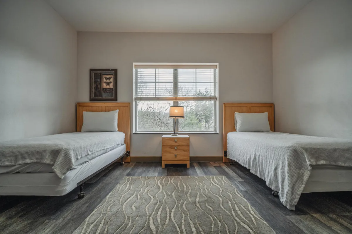 A serene bedroom featuring two twin beds separated by a central nightstand with a lamp and a large window letting in natural light. The space feels calm and is perfect for kids or friends.