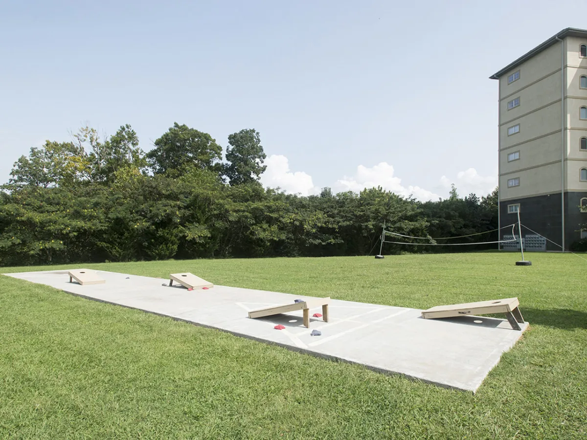 A vibrant outdoor recreation area featuring cornhole boards on a concrete pad, bordered by a lush green lawn and trees. A building can be seen in the background, implying easy access from the condos. The scene invites group play and socializing.
