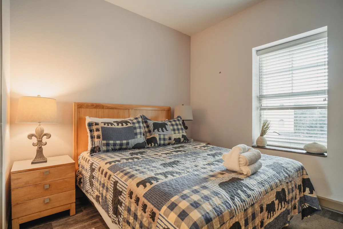 The image showcases a sunlit bedroom in a mountain-view condo, featuring a comfortable bed dressed with a patterned quilt, a nightstand with a glowing lamp, and a large window flooding the space with natural light. The scene exudes warmth and relaxation, perfectly blending inviting décor with the serenity of the Smoky Mountains just outside.