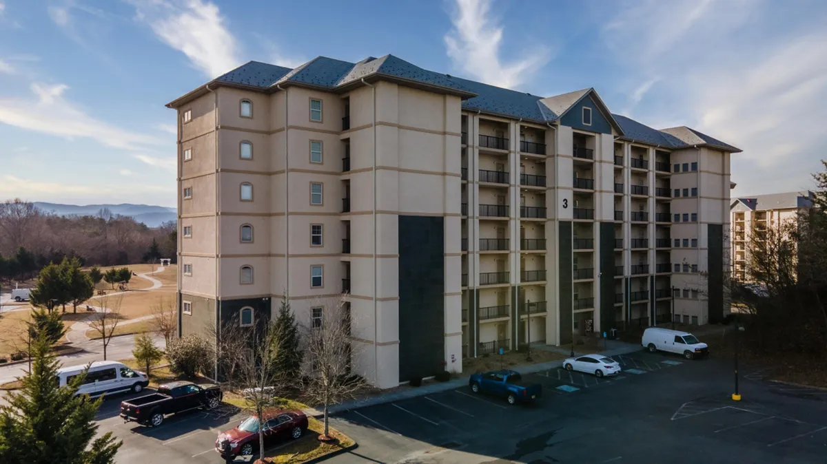 This image displays a multi-story condominium building surrounded by a spacious parking lot and lush greenery. The scene suggests a welcoming residential complex in a vibrant area, with easy access and a sense of community.