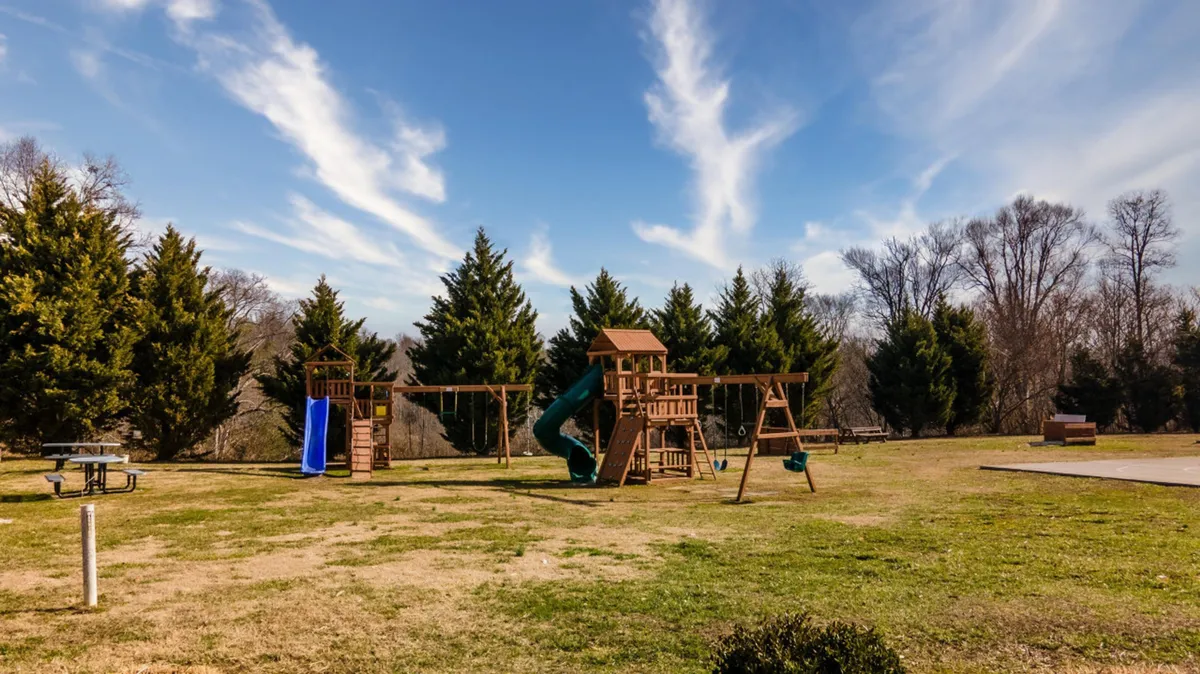 A vibrant playground surrounded by green grass, with picnic tables shaded by trees nearby. The scene is lively and inviting, suggesting a fun and safe environment for children and families to gather.