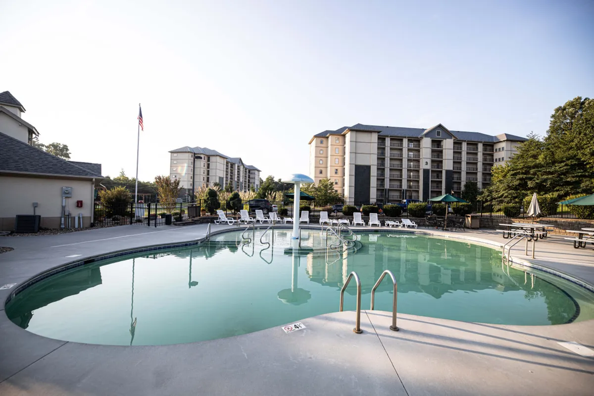 A sunny outdoor swimming pool surrounded by lounge chairs, with a playful fountain and nearby resort buildings visible in the background. Children can be seen splashing in the pool, suggesting a fun, family-friendly atmosphere.